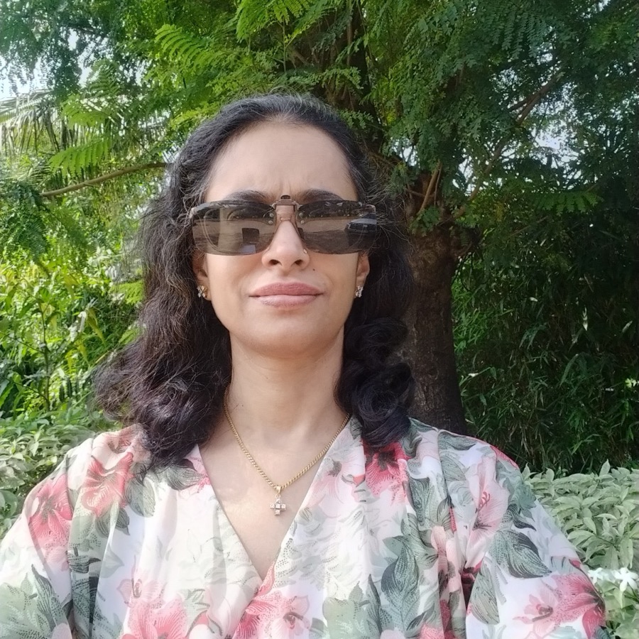 Photo of Priya Narayan, wearing sunglasses, with wavy dark hair, outdoors among greenery